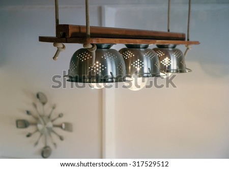 Warm lighting coming out from lamps inside beautiful utensils kitchen equipment, ropes and wood, hanged  from the ceiling Wall clock with forks and spoons on the background.