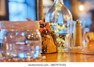 A warm, low-key shot featuring various glass vessels and a jar in the foreground, accented by twinkling fairy lights. The shallow depth of field draws focus to the textures and reflections on the glass, creating a cozy, festive Christmas atmosphere.