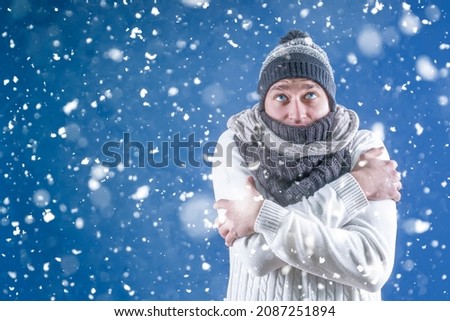 Warm. A man in a white sweater in a winter knitted hat and scarf stands under the falling snow. Snowfall. Winter. A person freezes in cold weather.