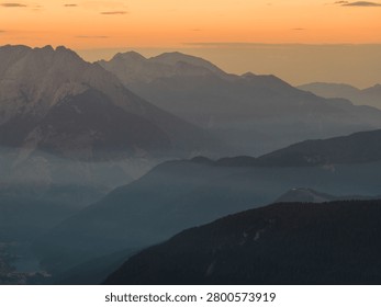 Warm orange sunset illuminates the dramatic peaks and valleys of the Dolomites, creating an atmospheric alpine landscape with layered mountain silhouettes in Italy.