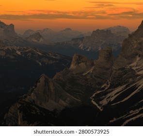 Warm orange sunset illuminates the dramatic peaks and valleys of the Dolomites, creating an atmospheric alpine landscape with layered mountain silhouettes in Italy.