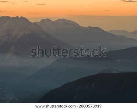 Warm orange sunset illuminates the dramatic peaks and valleys of the Dolomites, creating an atmospheric alpine landscape with layered mountain silhouettes in Italy.