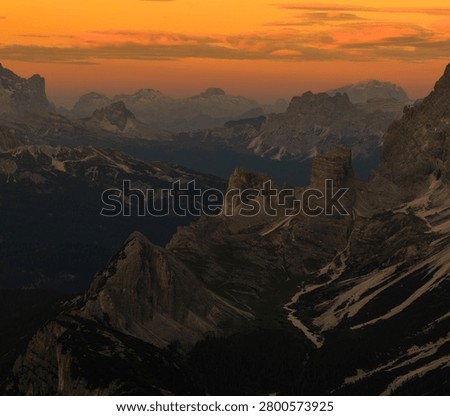 Warm orange sunset illuminates the dramatic peaks and valleys of the Dolomites, creating an atmospheric alpine landscape with layered mountain silhouettes in Italy.