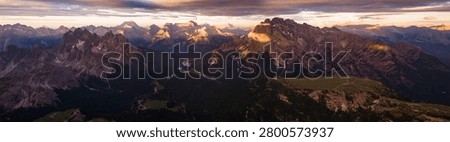 Warm orange sunset illuminates the dramatic peaks and valleys of the Dolomites, creating an atmospheric alpine landscape with layered mountain silhouettes in Italy.