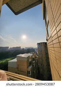 A warm orange sunset seen through a tall window framed by dark curtains, overlooking residential buildings and trees beneath dramatic evening clouds. The vertical composition creates a calm, reflective atmosphere suitable for travel, urban lifestyle, home interior, weather, and inspirational background concepts.