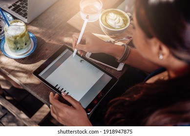 Warm Portrait Of A Asian Woman She Sits At Table With Coffee And Water Cups And Works On Her Tablet Outside In Sunny Day.