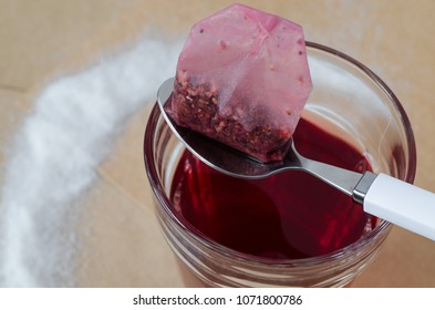 Warm Raspberry Tea In Glass And Tea Bag On White Spoon, Spilled Sugar Around, On Bright Brown Paper Background