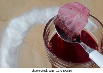 Warm Raspberry Tea In Glass And Tea Bag On White Spoon, Spilled Sugar Around, On Bright Brown Paper Background