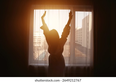 As the warm rays of the sun filter through the curtains, a woman stands by the window, stretching her arms joyfully.