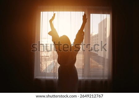 As the warm rays of the sun filter through the curtains, a woman stands by the window, stretching her arms joyfully.