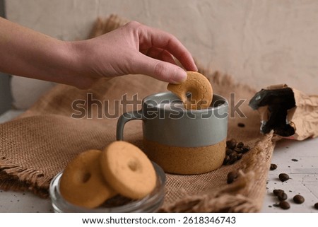 A warm, rustic coffee scene featuring a female hand holding a biscuit just above a mug of coffee, with cookies and scattered coffee beans on a surface and a woven fabric background