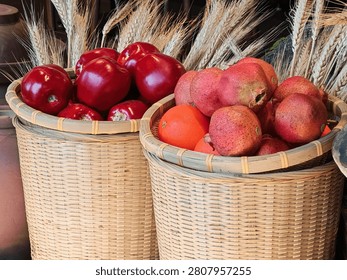 A warm rustic restaurant interior featuring a bamboo woven basket filled with fresh red apples and oranges, a central terracotta vase holding dried wheat stalks, and a wooden crate displaying green limes and oranges in layered arrangement. The decor evokes a harvest-rich, countryside charm.