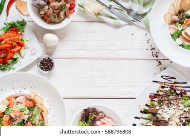 Warm salads frame on white wood flat lay. Top view on restaurant table with assortment of meat side dishes, free space. Buffet, banquet, menu concept