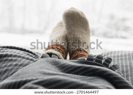 Warm socks and a soft blanket create a cozy atmosphere on a cold winter afternoon. Outside, snow gently falls, creating a peaceful winter scene.