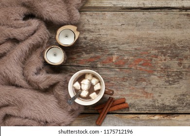 Warm Soft Blanket, Cup Of Hot Chocolate With Marshmallow, Candles And Cinnamon Sticks On Old Rustic Wood Background. Cozy Still Life. Top View Point.