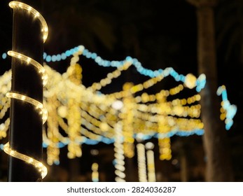 Warm spiral string lights wrap a dark pole in the foreground while softly blurred festive lights shimmer in the background at night.