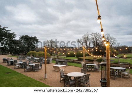 Warm string lights seen strewn above a large outside dining area at a country house and statley home in rural mid England.