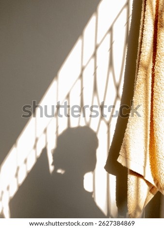 Warm sunlight creating a pattern of shadows from a window on a wall, with the shadow of a person and a soft towel visible. A simple yet evocative scene of light and domesticity