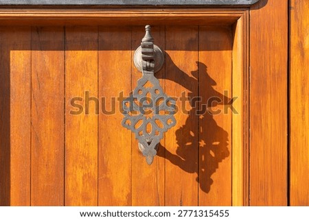 Warm sunlight glints off polished wooden door. Ornate metal knocker hangs with intricate patterns. Shadow of knocker dances on richly grained wood. Stillness suggests quiet anticipation or welcome