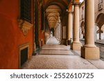 Warm sunlight illuminates the long, empty colonnade with its repetitive arches and columns, creating a striking perspective and highlighting the architectural beauty of bologna