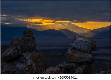 Warm sunset breaks through clouds illuminating distant mountains while rugged rocks in foreground frame tranquil valley below, creating mood of quiet awe and natural grandeur