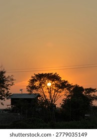 Warm sunset light behind trees and rural house silhouette during golden hour.
