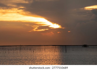 Warm sunset light breaks through clouds over a tranquil sea. Wooden stakes and a small offshore hut create a peaceful coastal scene with shimmering reflections and soft horizon tones.