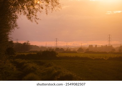 A warm sunset scene over fields in Bunny, Nottinghamshire, with golden light illuminating the rural landscape. Electricity pylons are visible in the distance, framed by trees and hedgerows. - Powered by Shutterstock