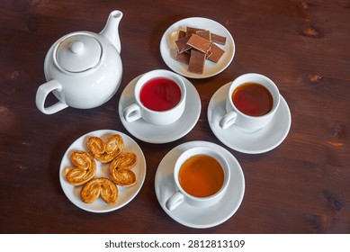 A warm tea time setup featuring a teapot, three cups of different teas (red, amber, and dark brown), chocolate bars, and golden palmiers on white porcelain plates against a rustic wooden table. The scene conveys relaxation and indulgence.