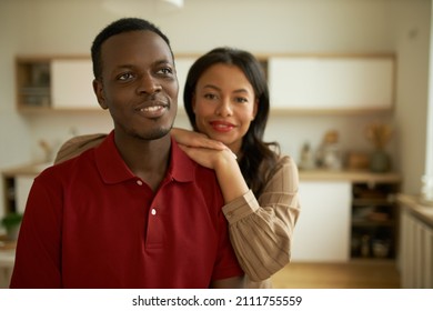 Warm tender relationships of attractive bearded dark-skinned male in red polo looking up dreaming, having his wife standing behind, leaning on his shoulder, showing her belief and dedication