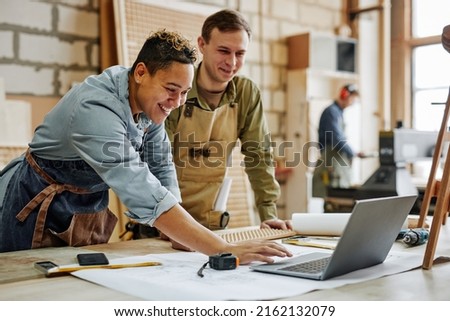 Warm toned portrait of animated carpenters using laptop and discussing furniture designs in workshop