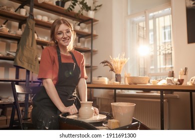 Warm toned portrait of young female artisan smiling at camera while working on pottery wheel in sunlit workshop and enjoying arts and crafts, copy space - Powered by Shutterstock