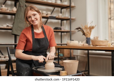 Warm toned portrait of young female potter smiling at camera while working on pottery wheel in workshop and enjoying arts and crafts, copy space