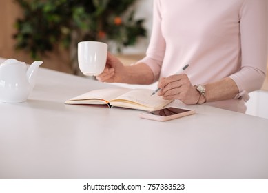 Warm Up. Close Up Of A Nice Senior Woman Making Notes And Drinking Tea While Standing In The Kitchen