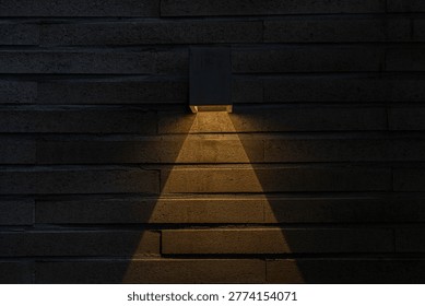 A warm wall light casts a triangular glow on a dark textured brick wall, creating a minimal architectural scene with dramatic contrast and atmosphere.