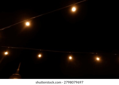Warm yellow bokeh string lights hanging on a wire against dark night background. Blurry light bulbs backdrop for romantic cafe, garden party, or festive decoration