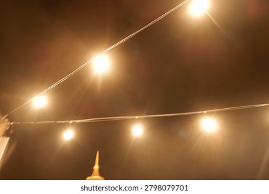 Warm yellow bokeh string lights hanging on a wire against dark night background. Blurry light bulbs backdrop for romantic cafe, garden party, or festive decoration