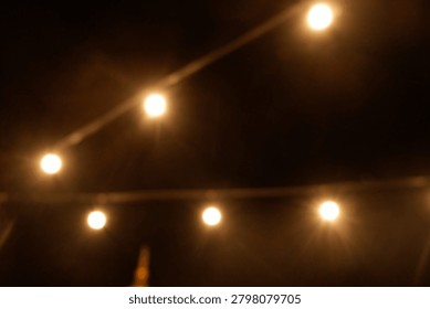 Warm yellow bokeh string lights hanging on a wire against dark night background. Blurry light bulbs backdrop for romantic cafe, garden party, or festive decoration