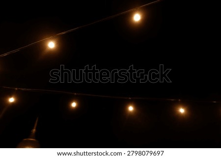 Warm yellow bokeh string lights hanging on a wire against dark night background. Blurry light bulbs backdrop for romantic cafe, garden party, or festive decoration