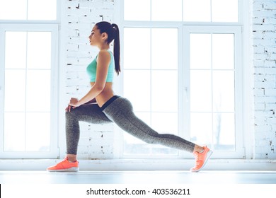 Warming up before training. Side view of young beautiful young woman in sportswear doing stretching while standing in front of window at gym