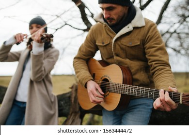 Warmly Dressed Bristled Man Playing Guitar Without Strap, Leaning Forward, Sitting On A Low Tree Branch. Next To Him A Woman, His Duet Partner Playing Violin. Over Autumn Countryside Landscape