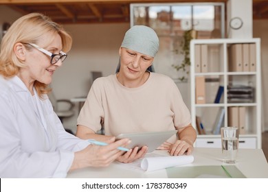 Warm-toned Portrait Of Mature Bald Woman Listening To Female Doctor Showing Info At Digital Tablet During Consultation On Alopecia And Cancer Recovery, Copy Space