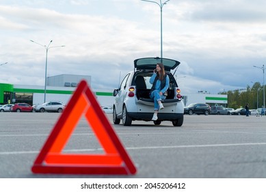 Warning red triangle. A white car with a flashing emergency light in the parking lot after an accident. Broken rearview mirror. Woman siting in trunk call insurance company or service repair and help.