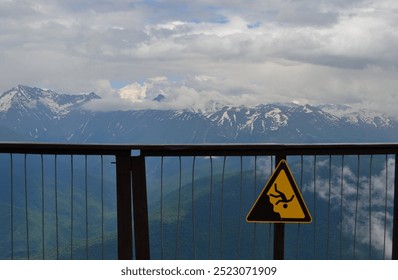 a warning sign against falling from a height in the mountains and a fence against the background of snowy peaks - Powered by Shutterstock