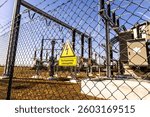 A warning sign for electrical current attached to a fence near a substation, emphasizing safety precautions around high-voltage areas.