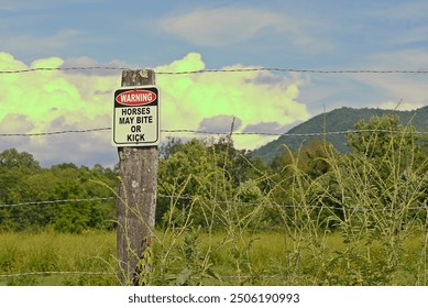 Warning sign that horse may bite or kick on a fence post with clouds and mountainous countryside in the background. - Powered by Shutterstock