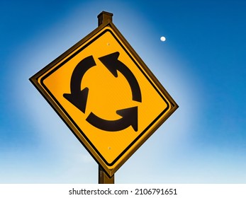 Warning sign with three counterclockwise arrows near a traffic roundabout (off camera), with waxing gibbous moon in the sky (selective focus on sign), for terrestrial and lunar motifs