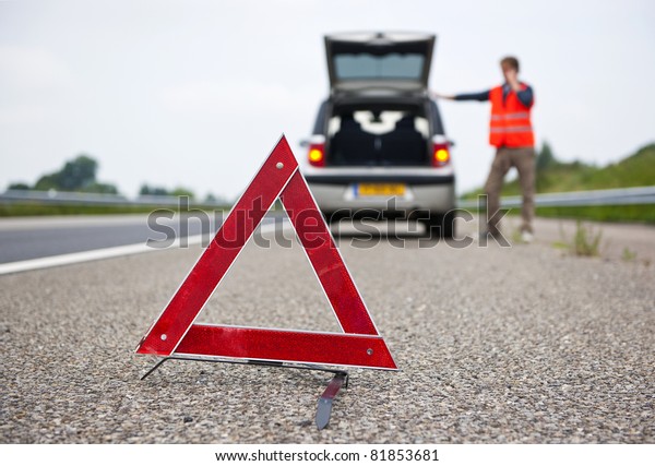 Warning triangle behind\
a broken down car with a motorist calling for assistance. Focus on\
the triangle