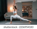 Warrior pose. Young woman with slim body type in white sportswear is indoors doing yoga.