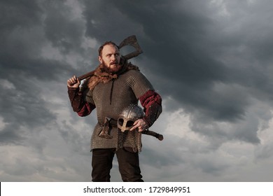 Warrior Viking in full arms with axe helmet and shield on dark sky background. Medieval scandinavian berserk red beard and fur costume attacks enemy. Concept historical photo